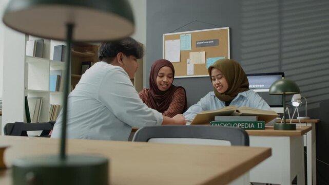 Asian Muslim female young adults and male classmate studying together in library, discussing project with encyclopedia. Perfect for university promo or diverse education campaigns