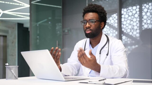 African American doctor talks on video call using laptop sitting at desk. Focused black man physician worker communicates remotely at online conference or has virtual medical consultation with patient