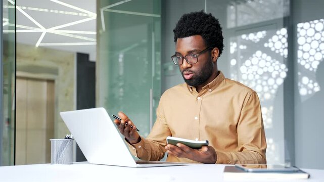 African American businessman sitting at desk in office, speaking during video call on laptop and taking notes. Professional black man manager asking questions or talking to client in workplace.