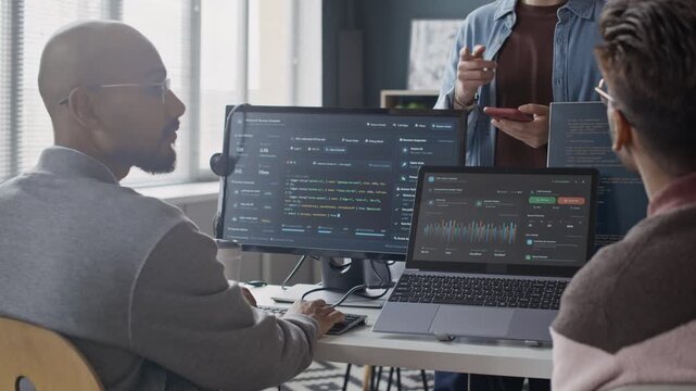 Slowmo shot of Multiethnic young adult developers collaborating in office, analyzing code on monitor and data charts on laptop. Clip is perfect for software development, IT consulting videos