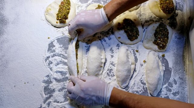 Gloved baker hands shaping filled dough with minced meat on a floured counter. Traditional Turkish pide or borek preparation in a bakery kitchen, showing handmade pastry production and culinary craft.