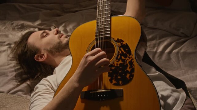Caucasian young man lying on bed, tuning acoustic guitar and strumming strings while creating new song. Footage is ideal for music education, creative hobby, or mental wellbeing marketing