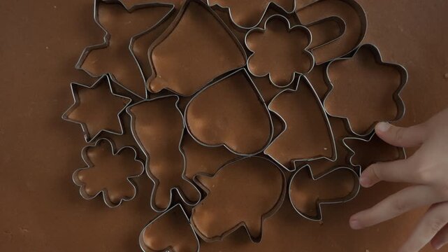 Mother and child place metal cookie cutters on gingerbread dough sheet in home kitchen. Family prepares sweet chocolate pastry shapes for holiday baking recipe food production process brown.