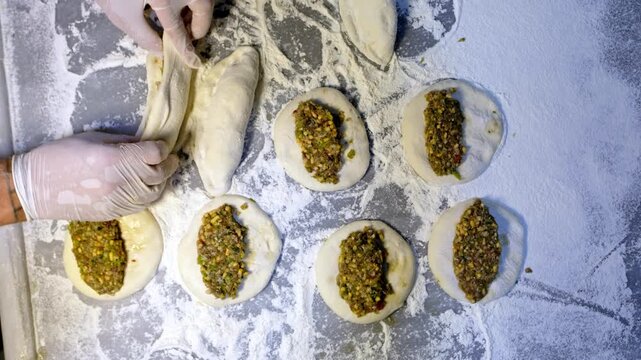 Gloved baker hands shaping filled dough with minced meat on a floured counter. Traditional Turkish pide or borek preparation in a bakery kitchen, showing handmade pastry production and culinary craft.