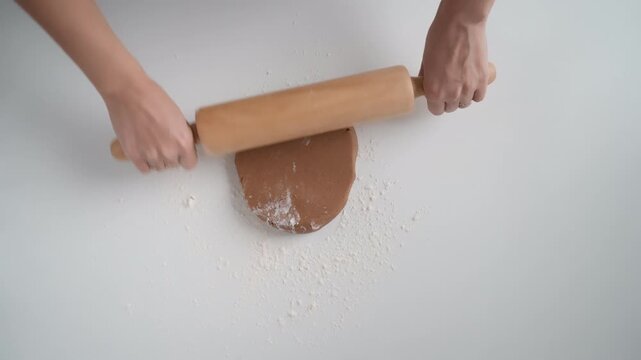 Woman hands roll out sweet gingerbread dough with wooden rolling pin on white table with flour. Female baker prepares homemade chocolate cookie pastry for baking in domestic kitchen room.
