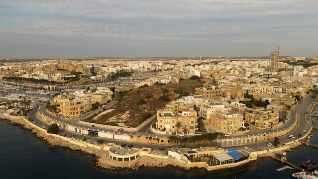 Aerial view of the coastal area featuring a marina with numerous boats, historic limestone buildings, and a winding waterfront road in Malta.
