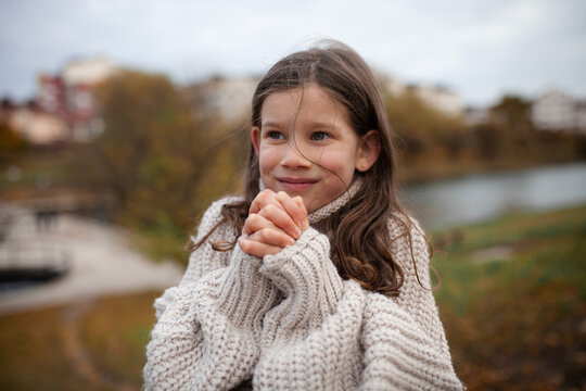Young girl feeling cold and warming her hands on an autumn day outdoors. Portrait of child with long dark hair wearing cozy oversized knit sweater, smiling.