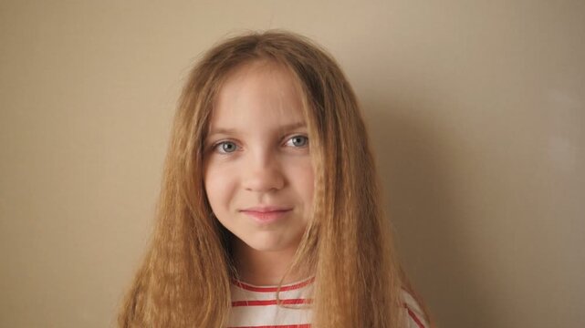 Portrait of exhausted female child looking into camera indoor. Small tired kid smiling against the background of beige wall. Little disappointed girl with long blonde hair inside. Close up