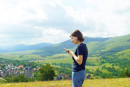 Woman viewing smartphone against mountain backdrop with settlements dispersed across green valley. Depicts access to information while traveling and technology integration in outdoor environments.