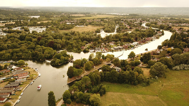 Aerial view of the Thames River winding through lush green countryside with residential houses, moored boats, and dense trees in Thames, United Kingdom.