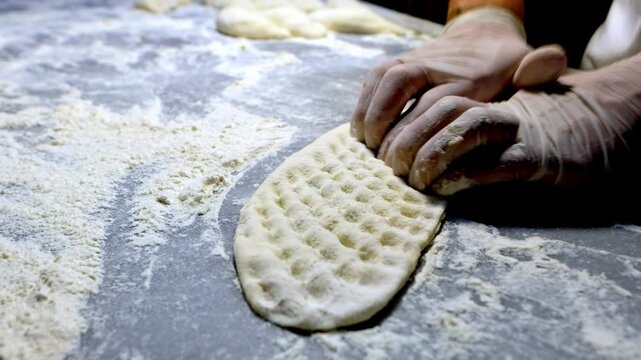 Close-up of gloved baker hands pressing and shaping pide dough on a floured counter. Traditional Turkish bakery work, handmade bread preparation, artisan kitchen craft and food production process.