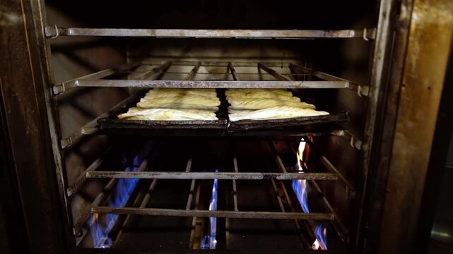 Pides baking on black trays inside an oven, with visible flames beneath. Bakery production, hot food preparation, baking process, professional kitchen workflow and restaurant production concept.