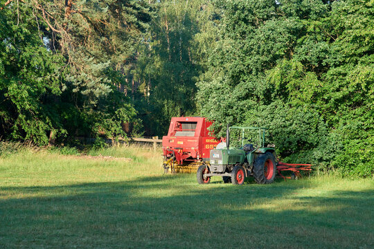Vintage green tractor with a red hay baler machine parked in a green grass field near a dense summer forest.