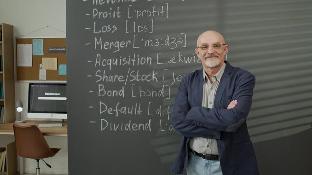 Portrait of middle aged Caucasian man standing with crossed arms in front of blackboard with business English vocabulary. Video is suitable for adult education and language school marketing