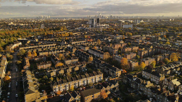 Aerial view of residential streets with rows of terraced houses, autumn trees, and the distant London skyline under a cloudy sky in London, England, United Kingdom.