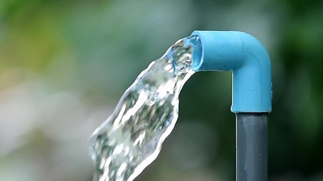 Clear water flowing from a blue pipe in a garden. The pipe is connected to a larger grey pipe and is angled to direct the water flow. The background is a blur of green leaves and soft sunlight.