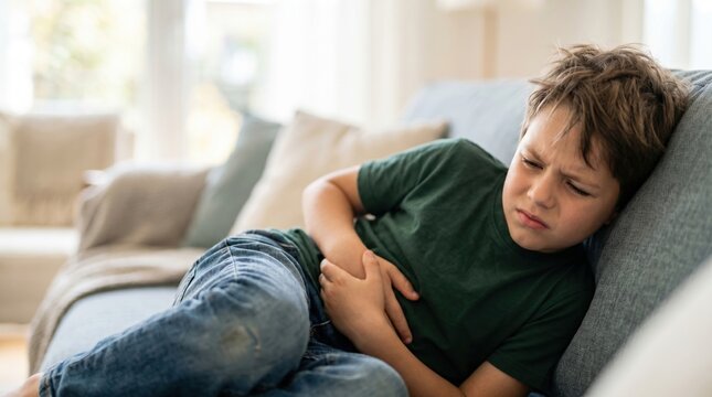 A boy is on a couch looking uncomfortable and holding his stomach. He appears to be in pain and is sitting in a bright living room with soft furniture