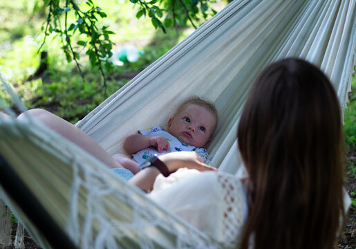 A young mother and baby relax in a white hammock in the garden on a sunny day. A cozy family morning outdoors. Mother-child bonding, mindful parenting, and relaxation.