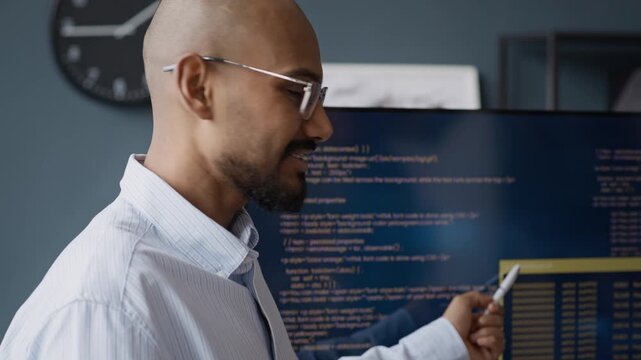 Tilt up shot of young Indian male developer pointing at monitor with code, explaining software programming during presentation in office. Video suitable for IT education and tech corporate marketing