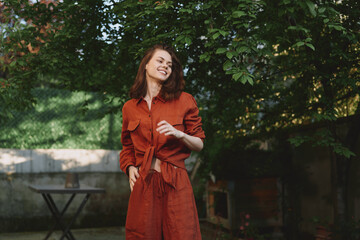 Obraz premium Smiling woman in a rust orange dress standing in a sunlit garden, casual pose and breeze in hair. Lifestyle portrait focusing on outdoor fashion and well being