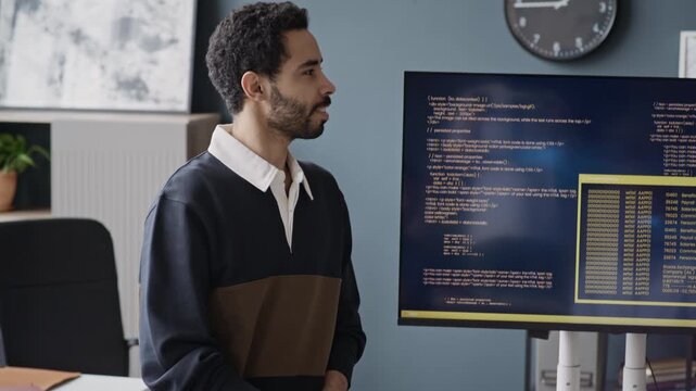 Handheld shot of young Middle Eastern male developer presenting software code on large monitor to colleagues in IT office. For tech startup marketing, software development, or corporate IT training