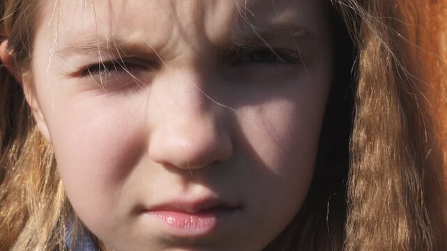 Female face of beautiful little girl illuminated by sun looking into camera outdoor. Portrait of small kid staring and squinting with a unhappy sight. Close up facial expression of serious child