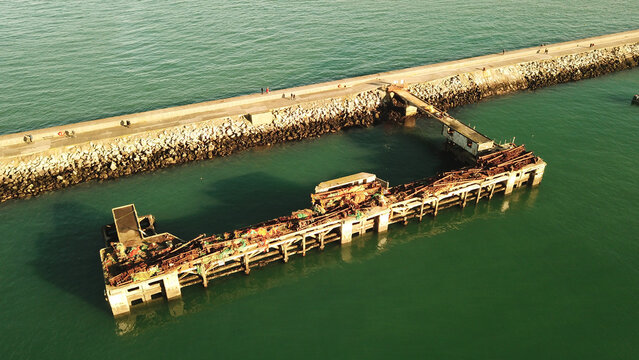 Aerial view of the historic breakwater and the weathered, rusted pier structure extending into the calm green sea in Brixham, England, United Kingdom.