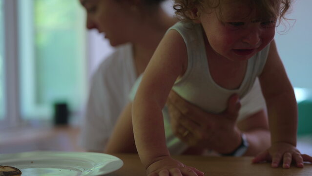 Frustrated toddler trying to climb onto table while mother holds child back during challenging parenting moment at home showing early childhood frustration and parental guidance