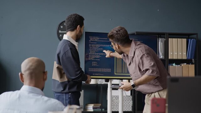 Handheld shot of motivated young multiethnic male programmers discussing code on screen during office meeting. Footage is suitable for software promo