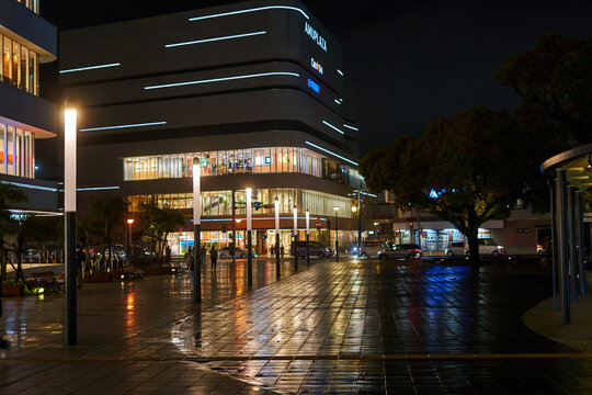 Night view of AMU PLAZA, a modern multi-story shopping complex in Japan, featuring distinctive horizontal blue LED accent lighting strips across its dark curved facade. The wet paved pedestrian