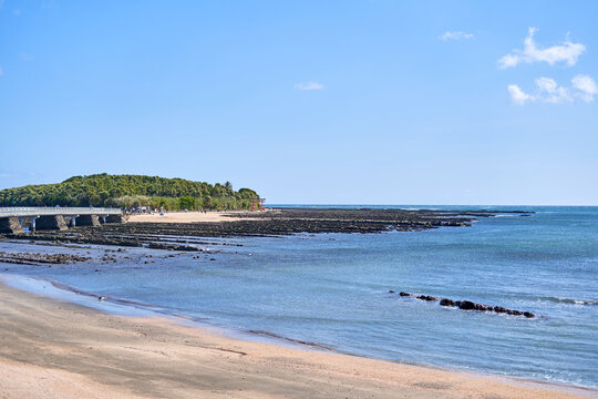 Scenic view of Aoshima Island in Miyazaki, Japan, featuring the iconic Yayoi Bridge connecting the small tropical island to the mainland. The foreground shows a sandy beach with gentle waves lapping
