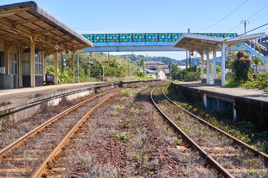 A rural or abandoned train station in Japan featuring two parallel railway tracks overgrown with grass and weeds, flanked by weathered concrete platforms with wooden shelters and canopies. A