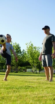 Father and son practicing soccer in park at sunset. Boy loses control of ball during juggling attempt while dad reacts with playful gesture, turning mistake into joyful family moment. Vertical