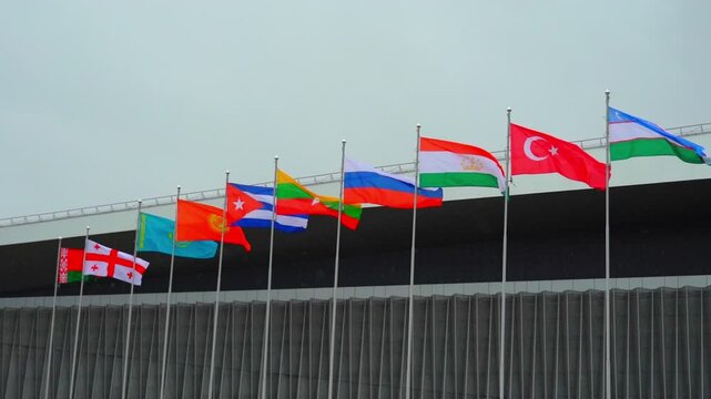 Multiple flags from various countries are displayed on tall flagpoles. The flags are seen fluttering in the wind against a cloudy sky. A mix of colors and designs can be observed as the flags represen
