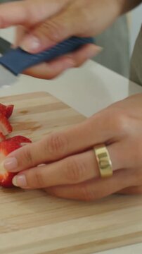 Vertical video: Gripping blue knife, cook in apron slicing strawberry at counter, preparing dish
