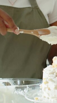 Vertical video: Lifting spoon, baker in olive apron scraping and decorating icing at counter, cake