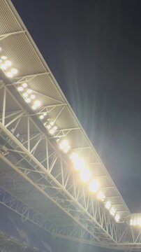 Football. Stadium floodlights illuminating the dark sky during a night game, showcasing powerful lamps on the roof structure and creating a bright, energetic ambiance for a sporting event