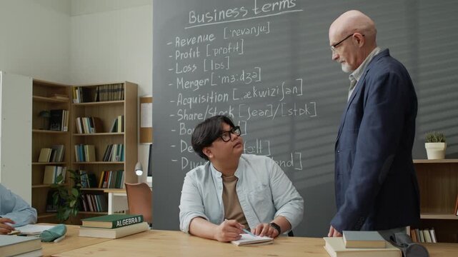 Senior Caucasian teacher explaining business terms to young adult Asian student writing in notebook. Video is perfect for marketing language schools and international education programs