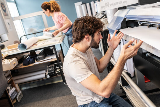A print shop worker is servicing a printer, carefully inspecting and cleaning internal components. The moment shows routine maintenance and attention to keeping production equipment in working order.