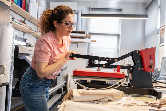 A print shop worker is operating a heat press machine for textile printing, carefully aligning the material before pressing. The scene shows a typical production step in custom garment printing with f