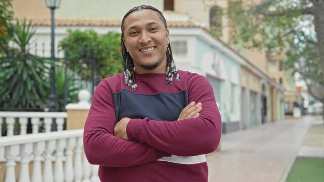 Man with beaded braids smiling with arms crossed on a street by white balustrade and storefronts; confidence approachability.