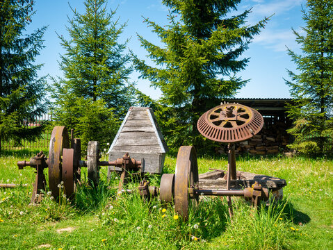 Rusty vintage farming equipment and iron wheels sit in a grassy meadow surrounded by evergreen trees under a bright blue sky.