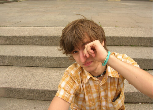Street city photo. Young white man with orange cotton checkered shirt, blue bracelet on hand, black coral beads on his neck sits on stair steps, rubs his eye, looks at camera. Concept of rest, relax