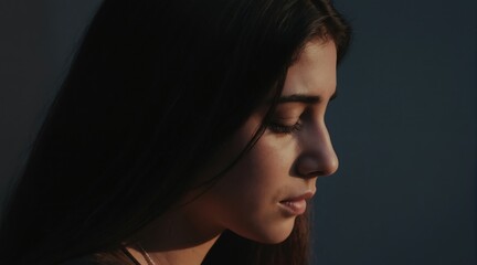 Fototapeta premium Young woman looks down with closed eyes in shadow. Soft light on face suggests sadness or deep thought. Her gaze is introspective, conveying quiet melancholy.