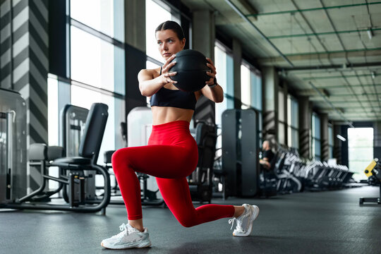 Athletic woman in red leggings doing forward lunge with medicine ball torso twist in gym