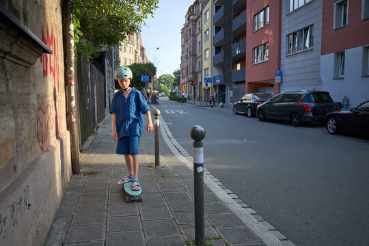 Young boy in blue outfit and helmet rides a skateboard down a city sidewalk