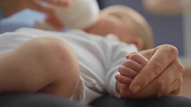 Newborn boy wearing a white onesie grasps adult finger and drinks from bottle in a nursery; tenderness bonding.