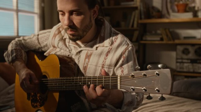 Caucasian young man playing acoustic guitar in home bedroom, focusing on fretboard. Footage ideal for music education, creative hobby, or online learning advertisements
