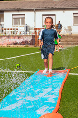 Obraz premium Child running on a colorful water slide with splashes of water in a sunny outdoor setting, surrounded by green grass and a residential background