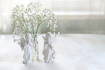 Obraz premium Beautiful composition with angel figurines and white gypsophila flowers in glass vase on table, abstract light background. symbol of faith, pray, believe, christianity. romantic, lovely atmosphere